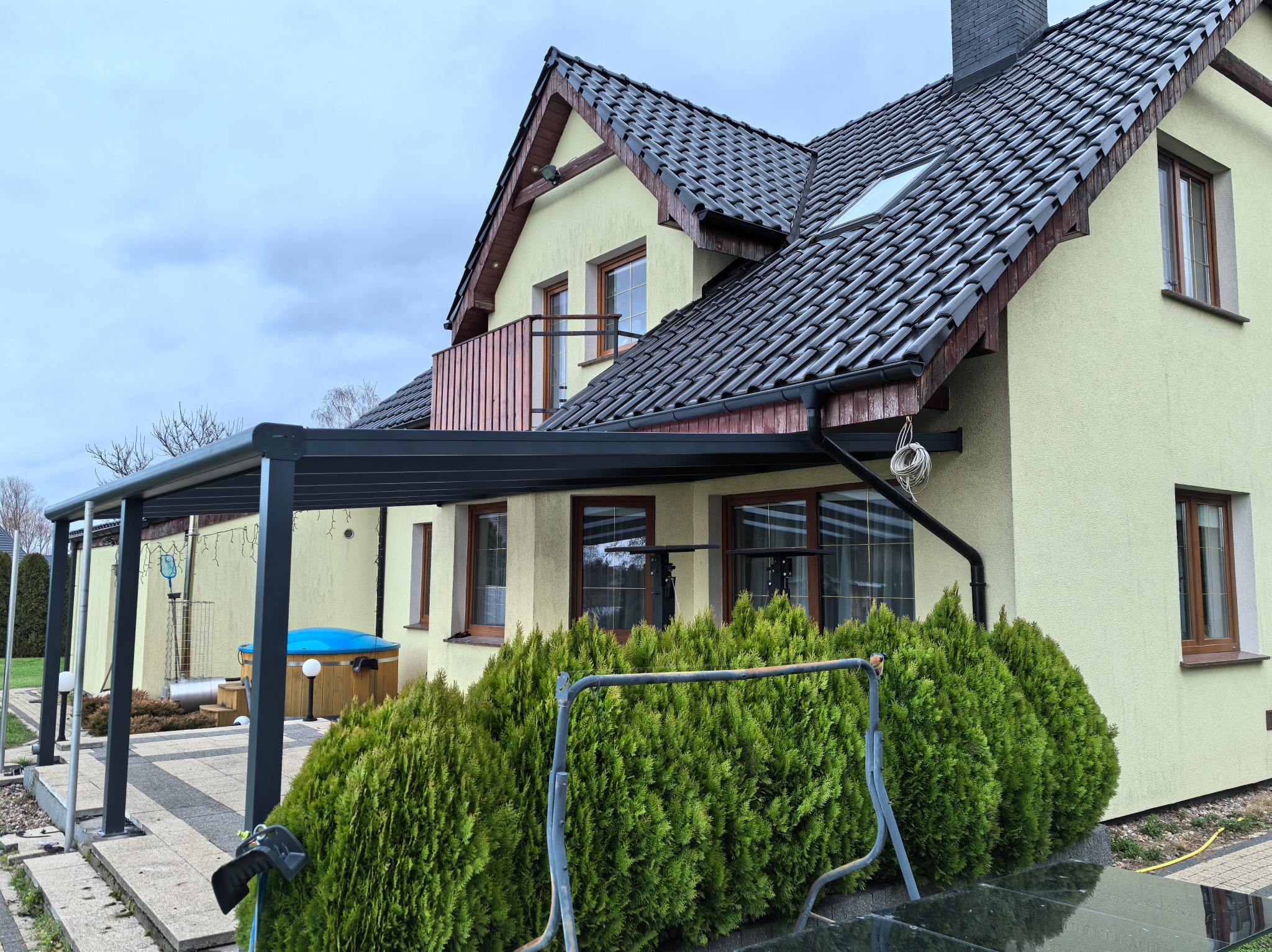 Czarna metalowa pergola przylegająca do jasnego domu z brązowymi oknami, na pierwszym planie rząd tuj, a w tle widoczna wanna spa z niebieską pokrywą.