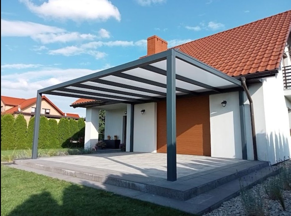 Nowoczesna pergola z metalową konstrukcją i przezroczystym zadaszeniem, przylegająca do białego domu z brązową roletą, otoczona zielonym trawnikiem i tujami.