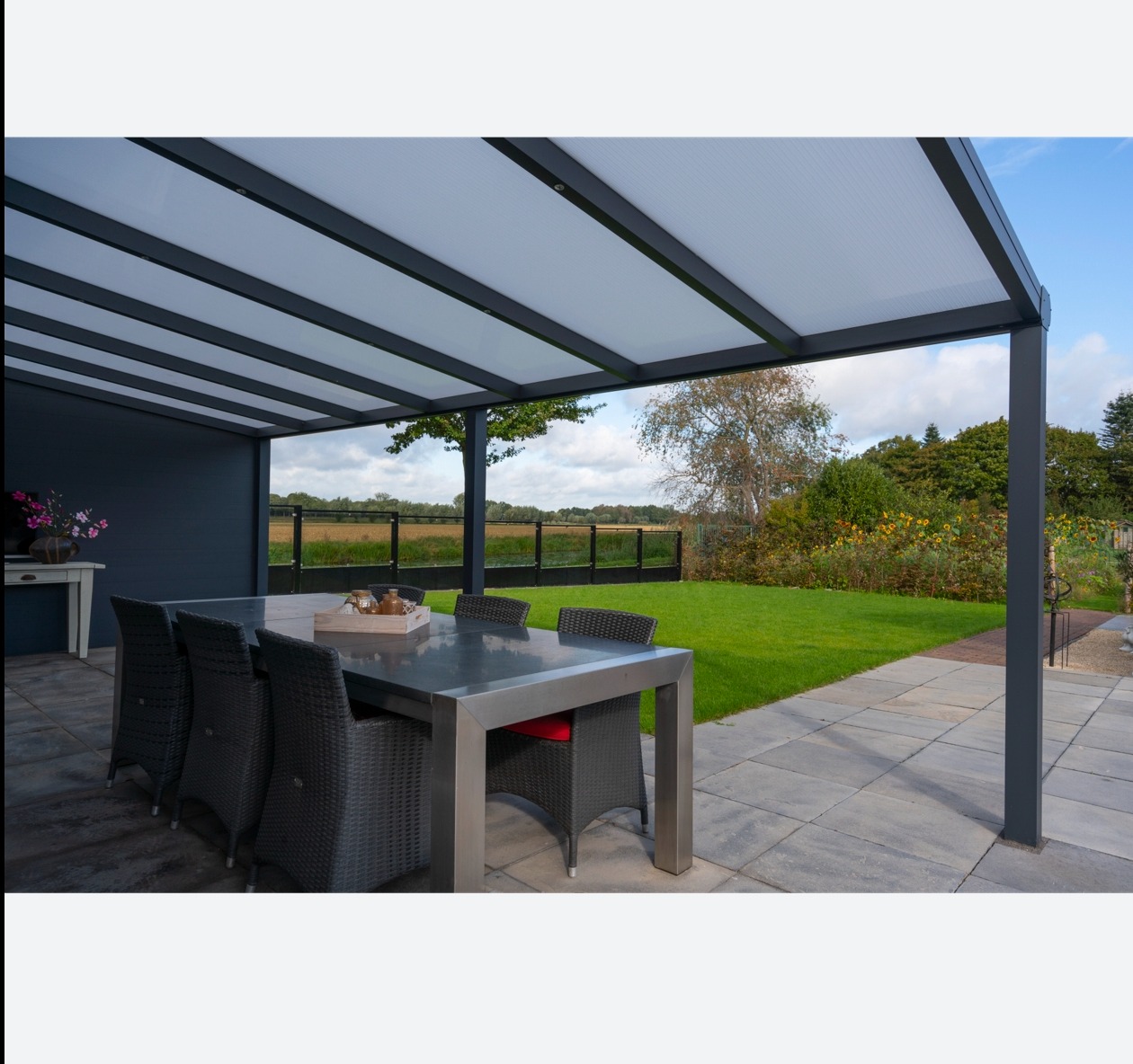 Nowoczesna, szara pergola z metalową konstrukcją i przezroczystym zadaszeniem, osłaniająca stół z krzesłami na patio z widokiem na zadbany trawnik i ogród.