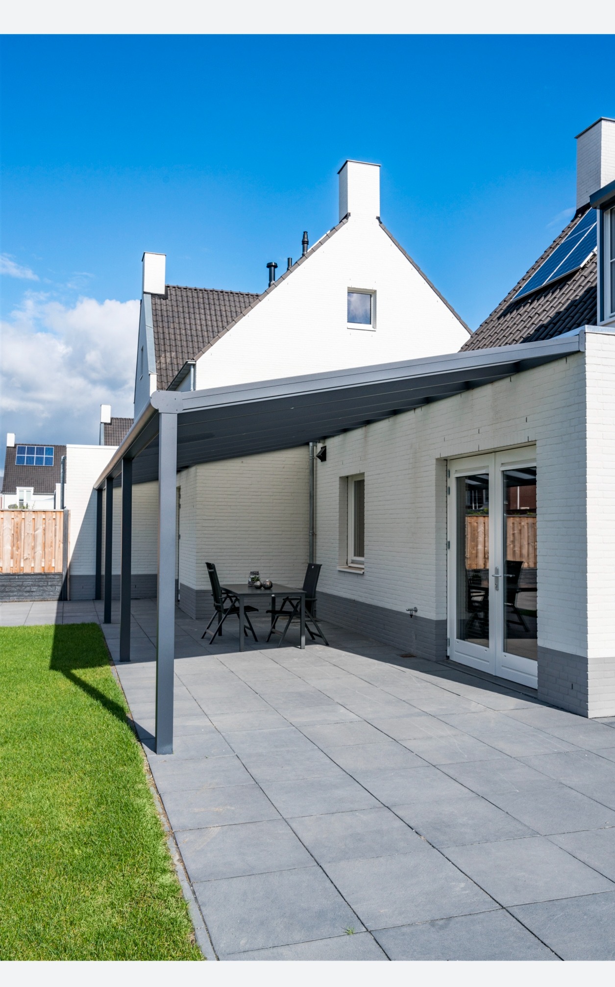 Nowoczesna pergola metalowa w kolorze grafitowym, przylegająca do białego domu z cegły, z widocznym zestawem mebli ogrodowych i zadbanym trawnikiem.