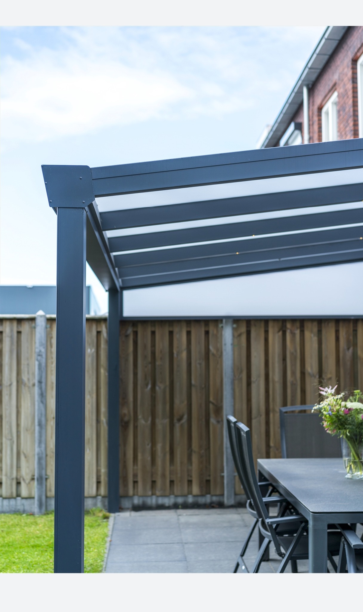 Nowoczesna pergola z metalową konstrukcją i lamelami, oświetlona punktowymi lampkami, z widocznym zestawem mebli ogrodowych i drewnianym ogrodzeniem w tle.