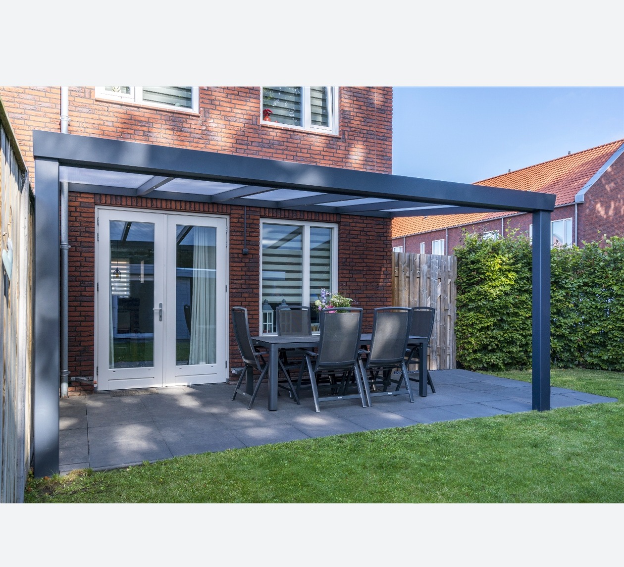 Nowoczesna, szara konstrukcja zadaszenia tarasu z przezroczystym dachem, stojąca na kostce brukowej obok trawnika, z meblami ogrodowymi i widokiem na ceglany dom.