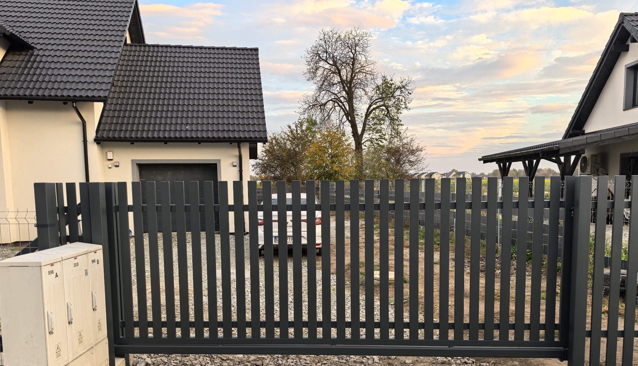 Nowoczesna, przesuwna brama w kolorze grafitowym z pionowymi sztachetami, widoczna z zewnątrz posesji, na tle domu z czarną dachówką i pochmurnego nieba o zachodzie słońca.