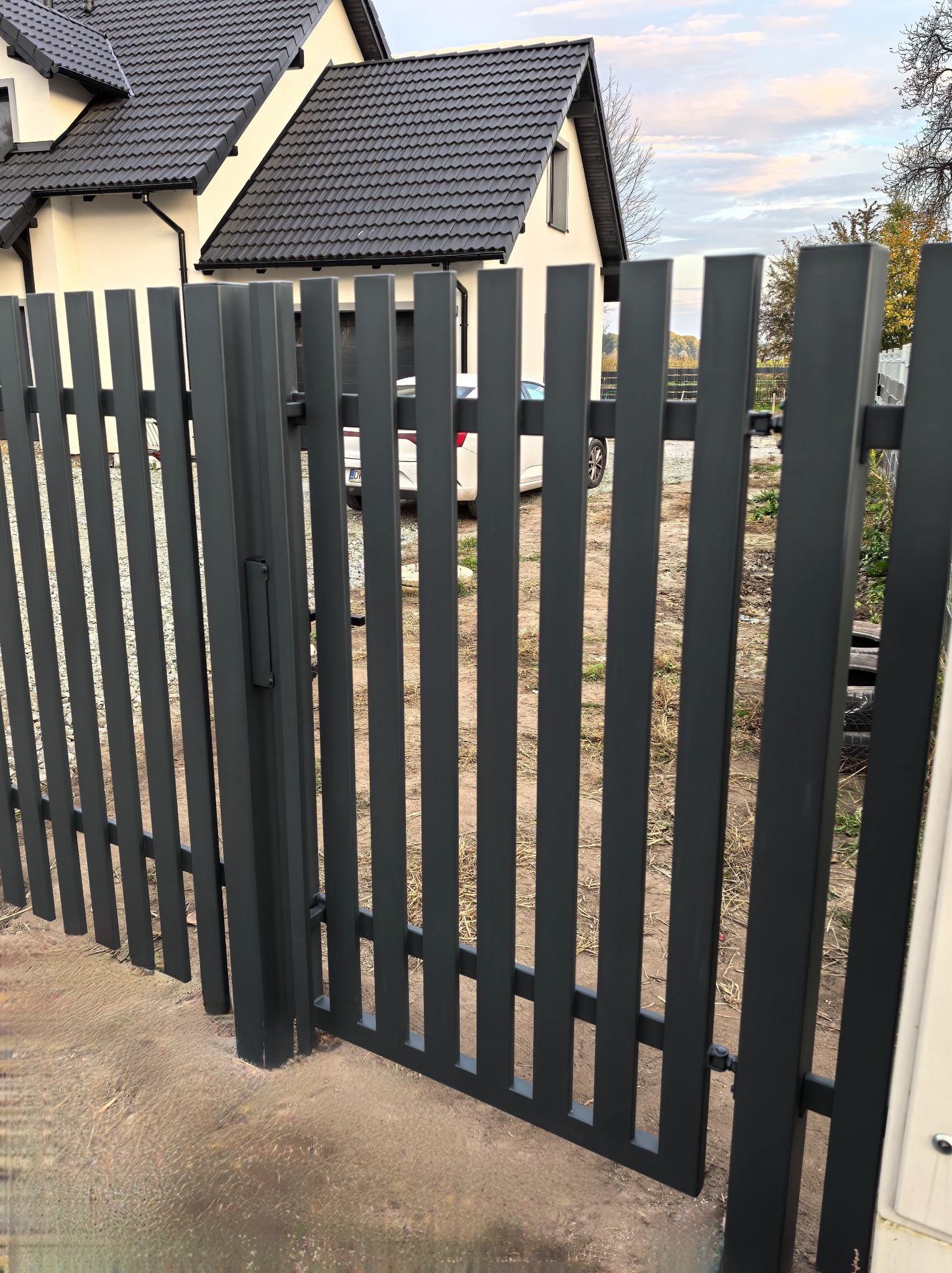 Nowoczesna, antracytowa brama posesyjna z pionowych profili stalowych, z widocznym zamkiem i fragmentem domu w tle.