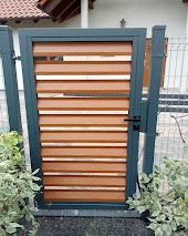 Nowoczesna furtka ogrodowa z poziomych paneli w kolorze drewna, obramowana ciemnoszarą ramą, osadzona między słupkami ogrodzeniowymi. Widoczne fragmenty siatki ogrodzeniowej i roślinność w tle.