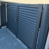 Nowoczesna, metalowa brama wjazdowa i furtka, pomalowane na grafitowo, z poziomymi, szerokimi lamelami, widoczne z bliskiej perspektywy na tle jasnej ściany budynku.