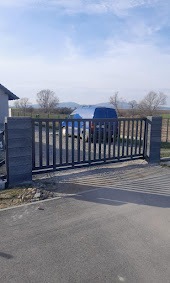 Nowoczesna, przesuwna brama metalowa z pionowymi szczebelkami, zamontowana między dwoma betonowymi słupkami o fakturze łupka. W tle widoczny samochód dostawczy i krajobraz z drzewami.