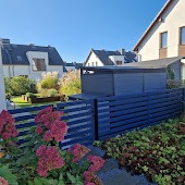 Nowoczesne ogrodzenie posesji z poziomych, ciemnych desek kompozytowych, z widocznym wejściem i fragmentem budynku w tle, otoczone zielenią i kwitnącymi krzewami.