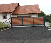 Przesuwna brama wjazdowa z poziomymi panelami w kolorze drewna, osadzona na czarnej, żwirowej nawierzchni podjazdu, widoczna na tle domu z czerwoną dachówką.