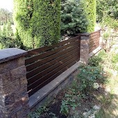 Nowoczesne ogrodzenie panelowe z brązowych, poziomych desek, osadzone na kamiennych słupkach z wyraźną fakturą, otoczone zielenią w przydomowym ogrodzie.