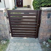 Nowoczesna, brązowa furtka z poziomych desek zamontowana między kamiennymi słupkami, prowadząca do domu z ciemnymi drzwiami wejściowymi i zielenią w tle.