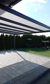 Nowoczesna pergola metalowa z białym zadaszeniem, rzucająca cień na szary brukowany taras, w tle zielony żywopłot i huśtawka ogrodowa.