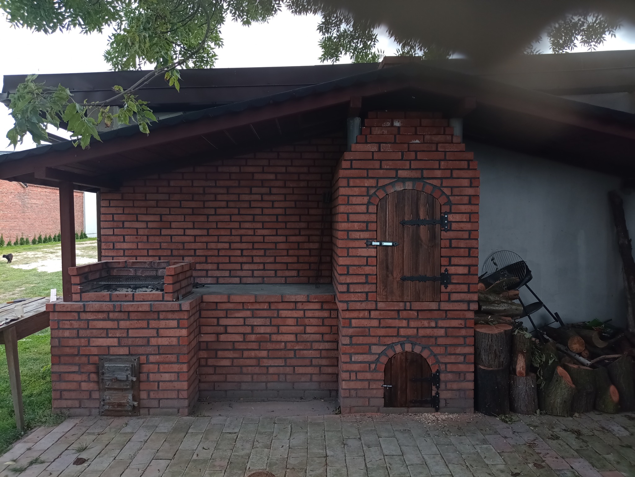 Murowana kuchnia letnia z grillem i wędzarnią, z drewnianymi drzwiczkami, usytuowana na brukowanym patio, z widocznymi zapasami drewna obok.