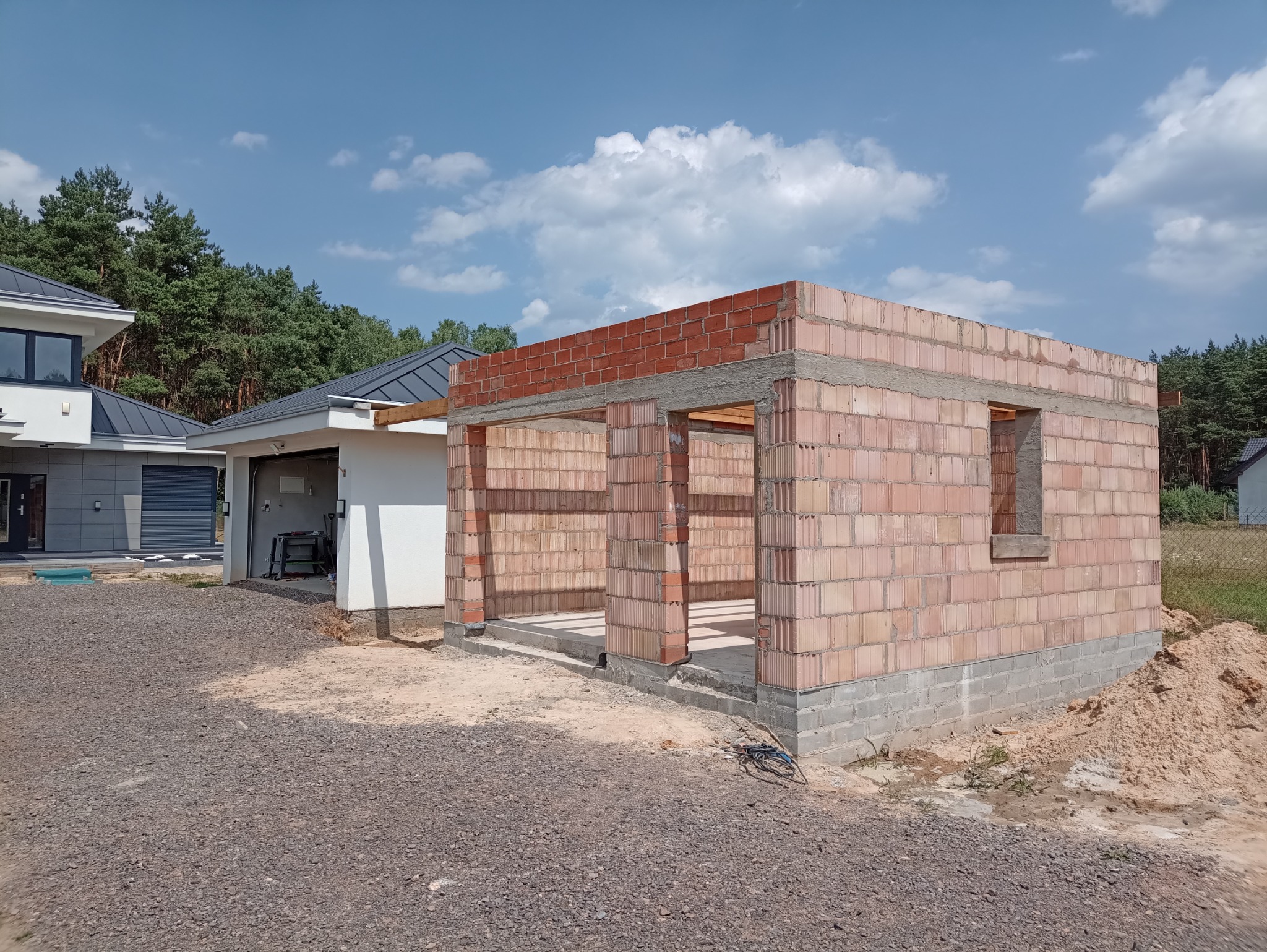 Surowy stan budynku z czerwonej cegły, widok z zewnątrz, z widocznymi otworami okiennymi i wejściowymi, na tle błękitnego nieba z chmurami i fragmentem sąsiedniego domu z garażem.