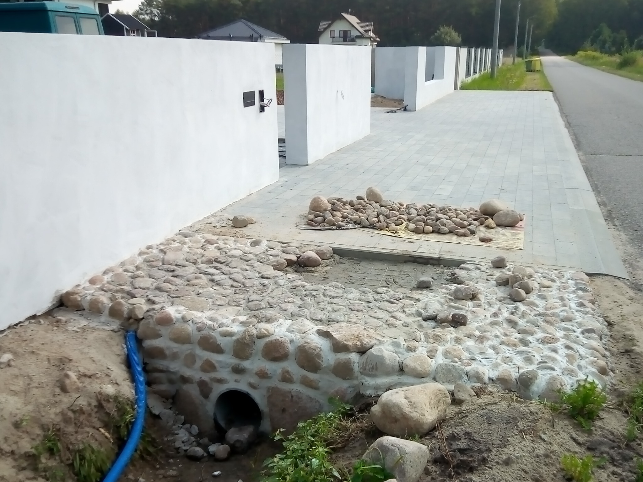 Wykończony drenaż z kamieni polnych przy wjeździe na posesję, widoczna rura odprowadzająca wodę i fragment białego muru.