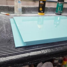 MEB-LAK - Turkusowy front meblowy z frezowanym uchwytem, leżący na warsztatowym stole. Widoczne preparaty do polerowania i wykończenia. Błyszcząca powierzchnia.