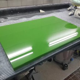 MEB-LAK - Błyszczący, zielony front meblowy leży na blacie roboczym. Widoczne odbicie światła, szlifierka i odkurzacz. Detal z produkcji mebli na wymiar.