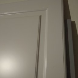 MEB-LAK - Fragment białej szafy na wymiar z frezowanym frontem, widoczny bok i ściana w tle. Ujęcie z bliska, podkreślające detale wykończenia.