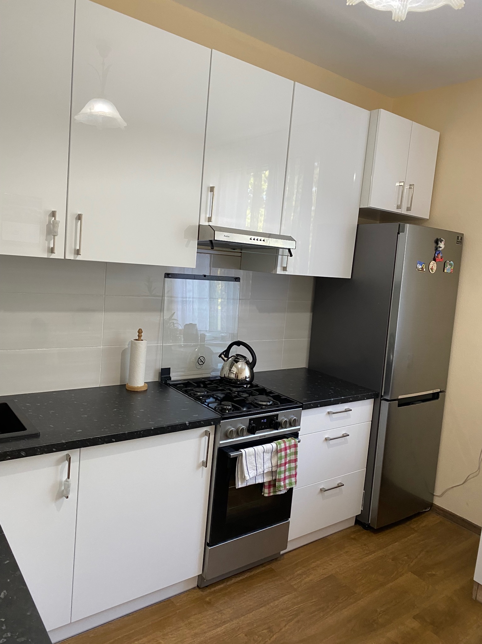 Biała kuchnia z czarnym blatem, płyta gazowa z czajnikiem, srebrna lodówka z magnesami, jasne ściany. Widok na szafki wiszące i stojące.