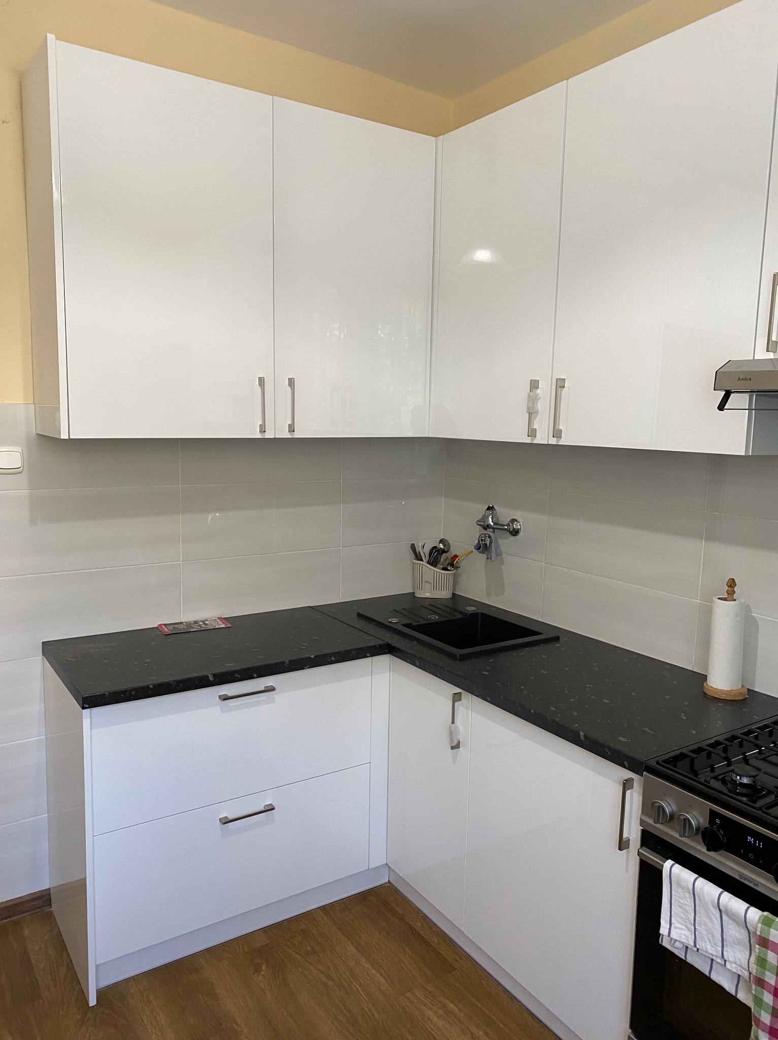 Biała, narożna kuchnia z połyskiem i czarnym blatem. Widoczny zlew, bateria i płyta gazowa. Na blacie przybory kuchenne i ręcznik papierowy.