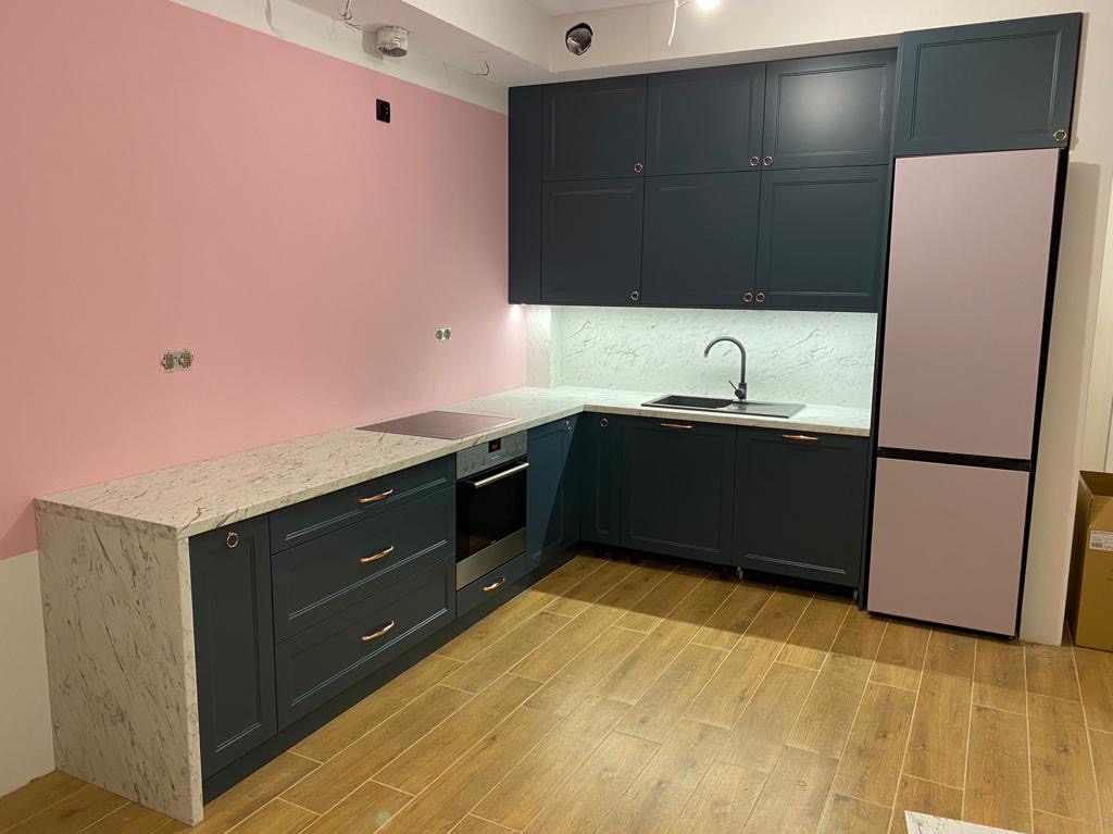 Nowoczesna kuchnia z ciemnymi szafkami, marmurowym blatem i różową lodówką, oświetlona ledami. Uwagę zwracają miedziane uchwyty i różowa ściana w tle.