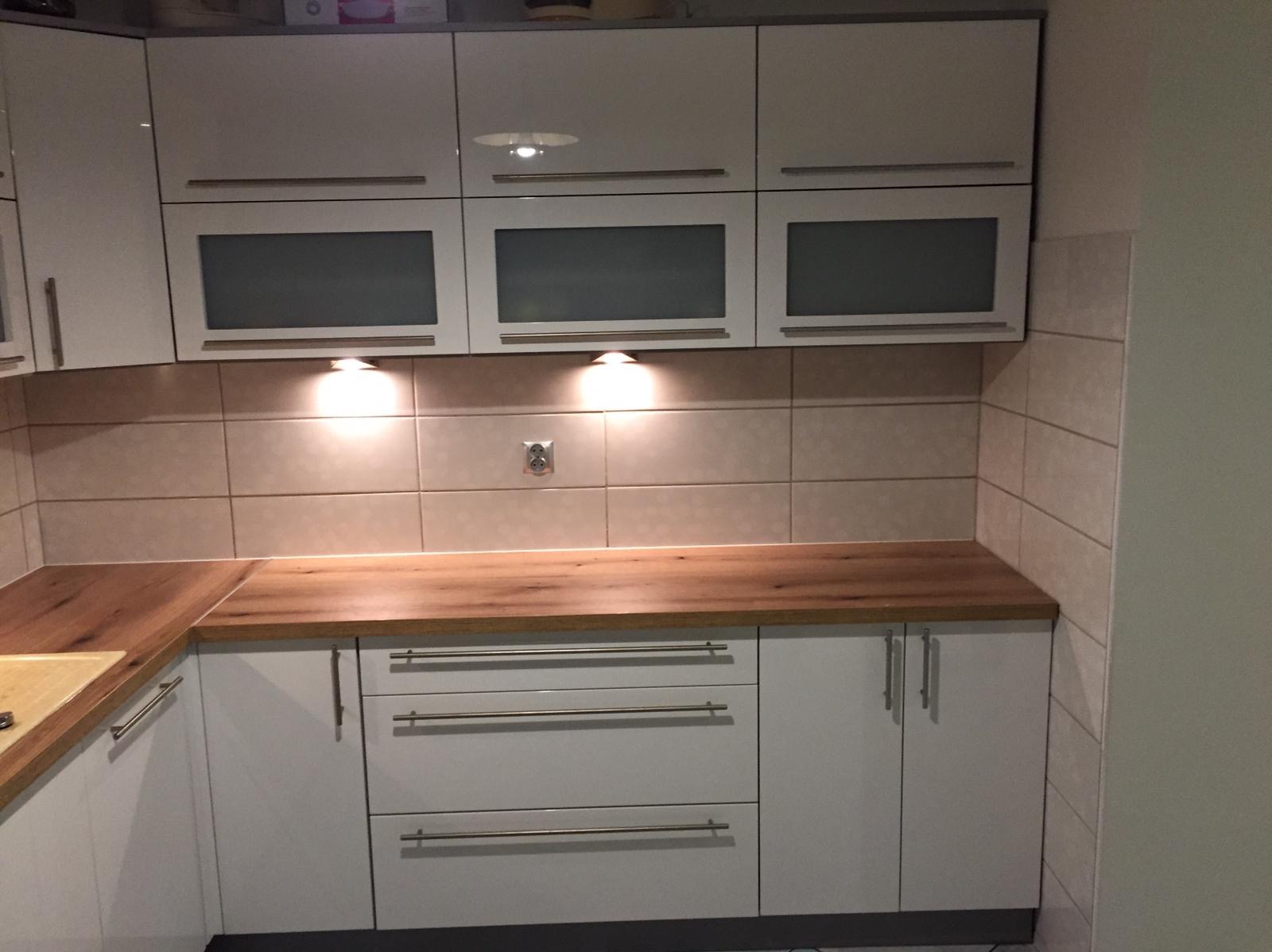 Biała kuchnia z drewnianym blatem i szklanymi szafkami wiszącymi, oświetlona punktowo, z beżowymi płytkami na ścianie i widocznym gniazdkiem elektrycznym.