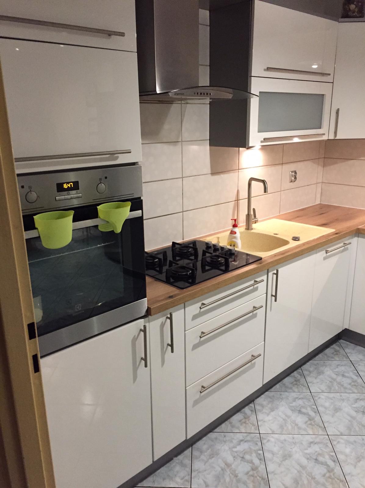 Biała kuchnia z drewnianym blatem, z piekarnikiem z zielonymi akcesoriami, czarną płytą grzewczą i beżowym zlewem. Uwagę zwracają minimalistyczne uchwyty szafek.