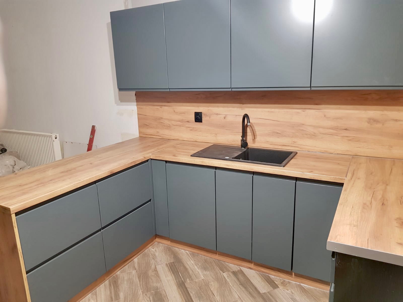 Szare, matowe szafki kuchenne z drewnianym blatem i panelem. Widoczny zlew i bateria. Częściowo wykończone pomieszczenie. Nowoczesna kuchnia narożna.