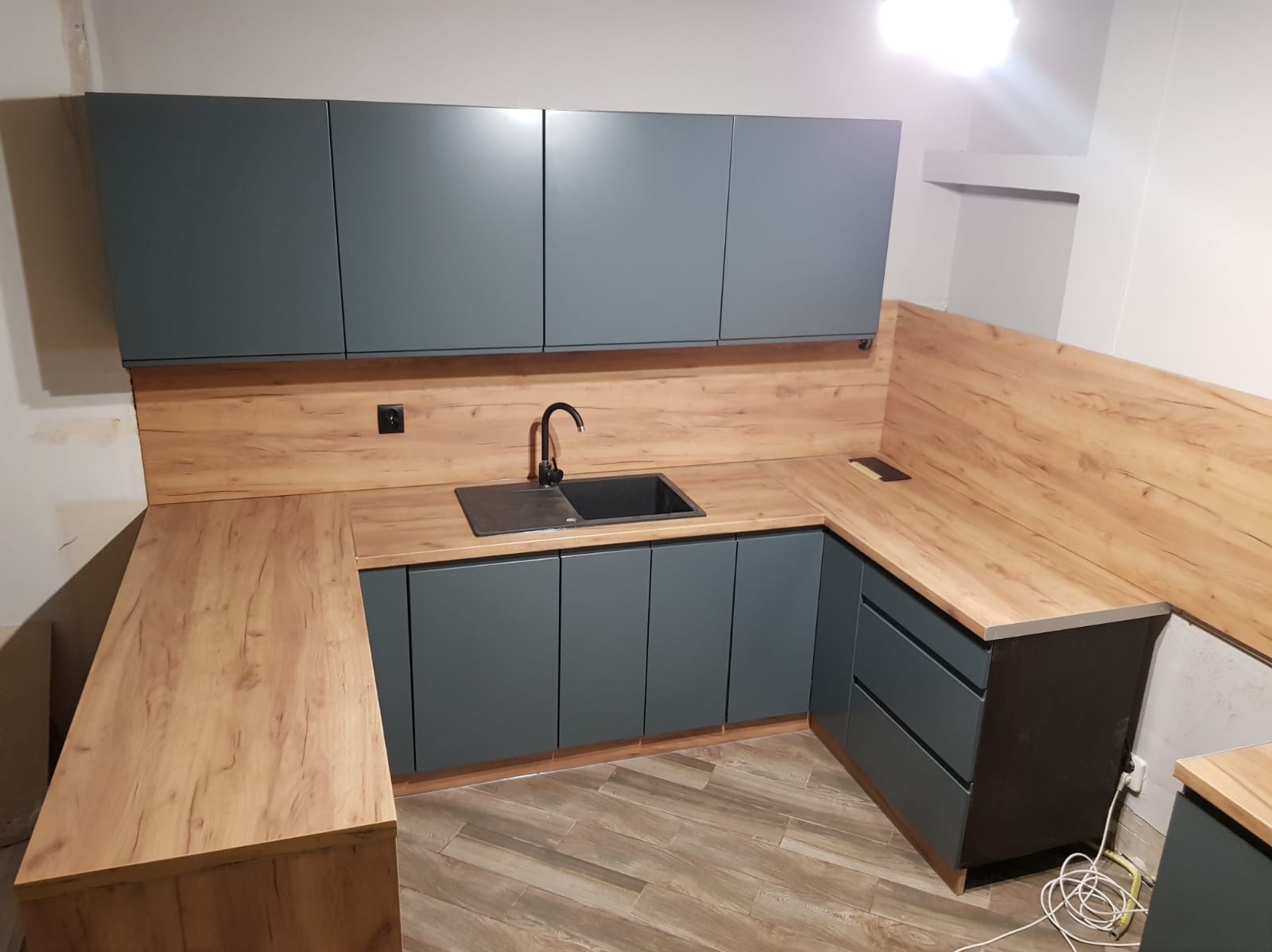 Szara zabudowa kuchenna z drewnianym blatem i panelem ściennym. Umywalka wpuszczona w blat. Widoczny fragment podłogi z płytek imitujących drewno.