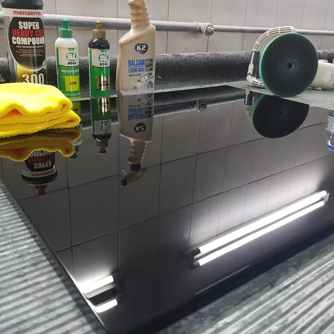 Błyszczący, czarny blat z odbiciami lamp, środków do polerowania i ściereczki. Widoczne płytki na ścianie i podłodze. Detale wykończenia blatu.