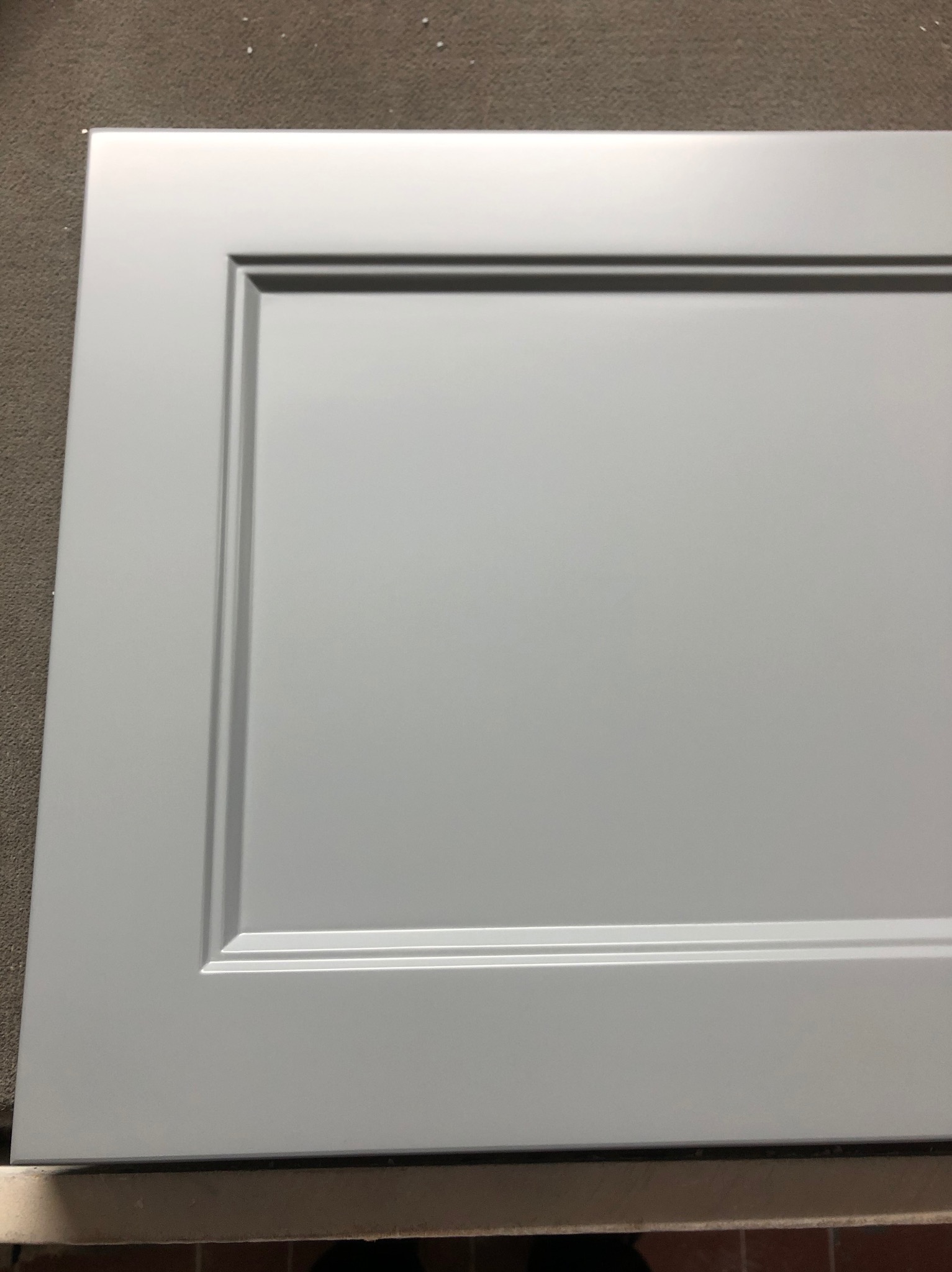 Szary front meblowy z frezowanym obramowaniem, leżący na szorstkiej powierzchni. Detal wykończenia szafki lub szuflady w zbliżeniu.