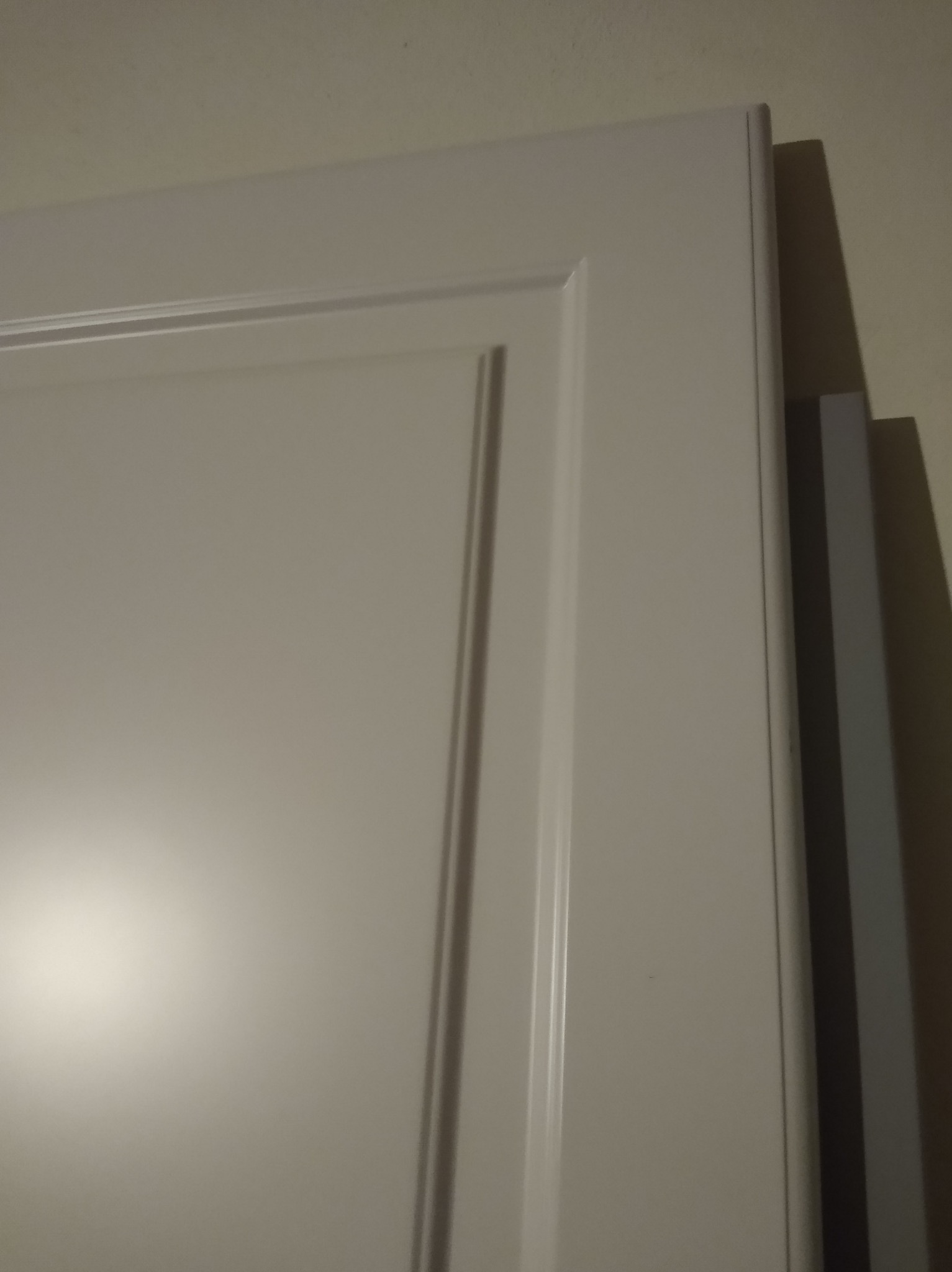 Fragment białej szafy na wymiar z frezowanym frontem, widoczny bok i ściana w tle. Ujęcie z bliska, podkreślające detale wykończenia.