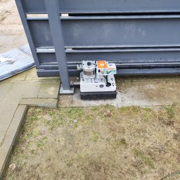 ML AUTOMATYK MAREK LIS - Szara brama przesuwna z zamontowanym automatem, widoczne przewody i elementy sterujące, obok leży materac, a w tle trawa i kostka brukowa.