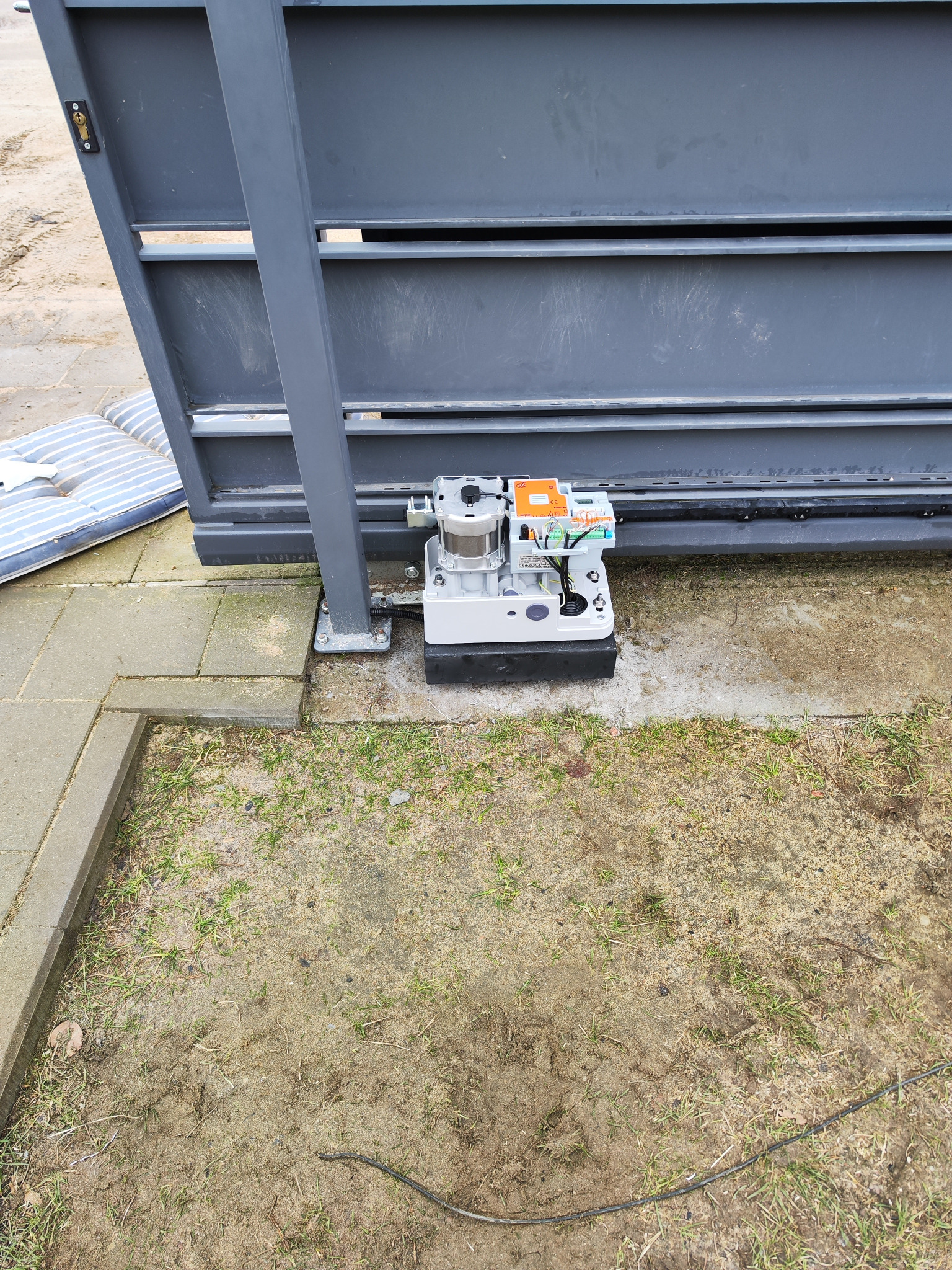Szara brama przesuwna z zamontowanym automatem, widoczne przewody i elementy sterujące, obok leży materac, a w tle trawa i kostka brukowa.