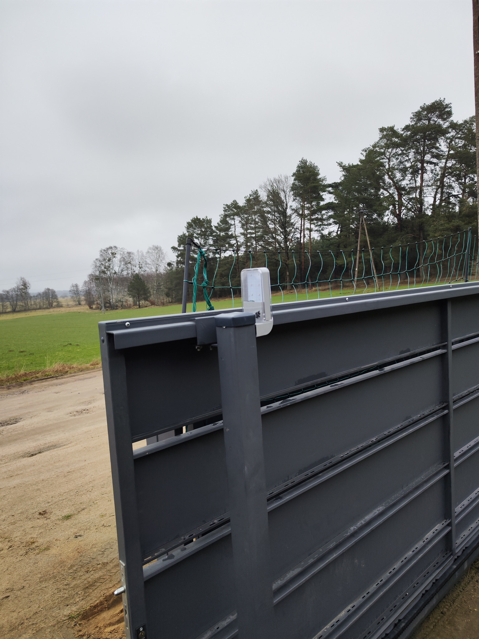 Szara brama wjazdowa z zamontowanym czujnikiem ruchu, widok z dołu na tle zielonego pola i drzew pod pochmurnym niebem.