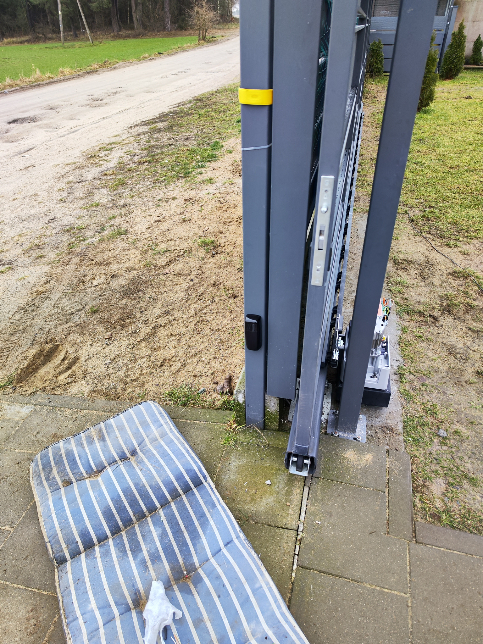 Szara brama przesuwna z widocznym napędem i elementami automatyki, obok leży brudny materac w niebieskie paski, a w tle widoczna droga gruntowa.