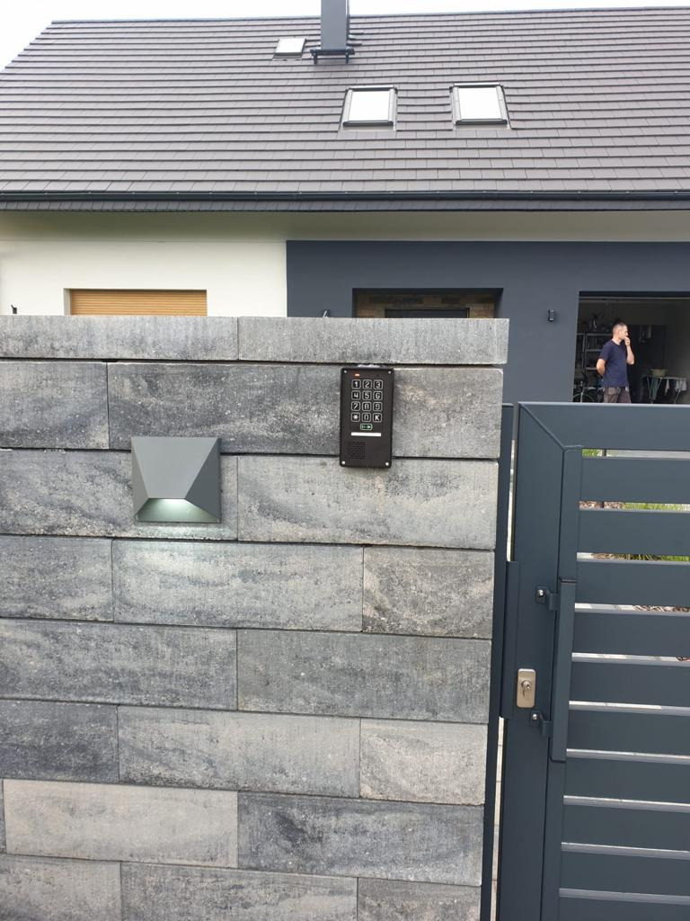 Szary, kamienny mur z zamontowanym panelem numerycznym domofonu i nowoczesną lampą elewacyjną, widoczna brama wjazdowa i fragment domu z szarą dachówką w tle.