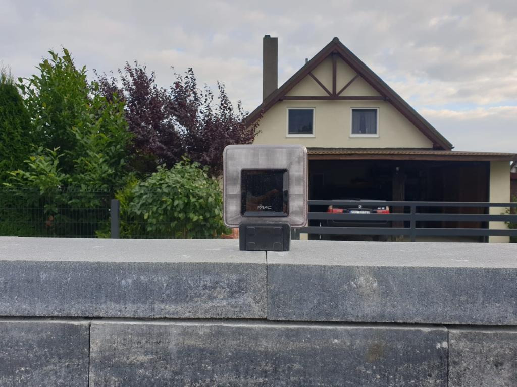 Fotokomórka FAAC zamontowana na szarym, kamiennym słupku bramy wjazdowej, na tle domu z garażem i zielenią.