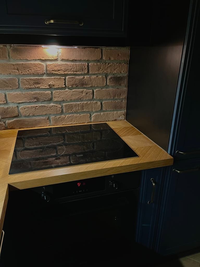 Fragment kuchni z indukcyjną płytą grzewczą, blatem z jasnego drewna i ścianą z cegieł, oświetloną punktowym światłem, w zabudowie z ciemnymi, granatowymi szafkami.