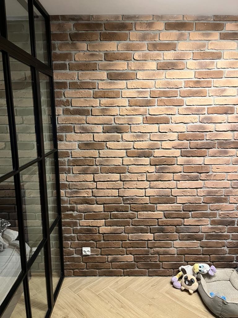 Ściana z dekoracyjnej cegły w Luboniu, z widocznym gniazdkiem elektrycznym i fragmentem czarnej ramy przeszklenia, uzupełnona o psie akcesoria na podłodze.