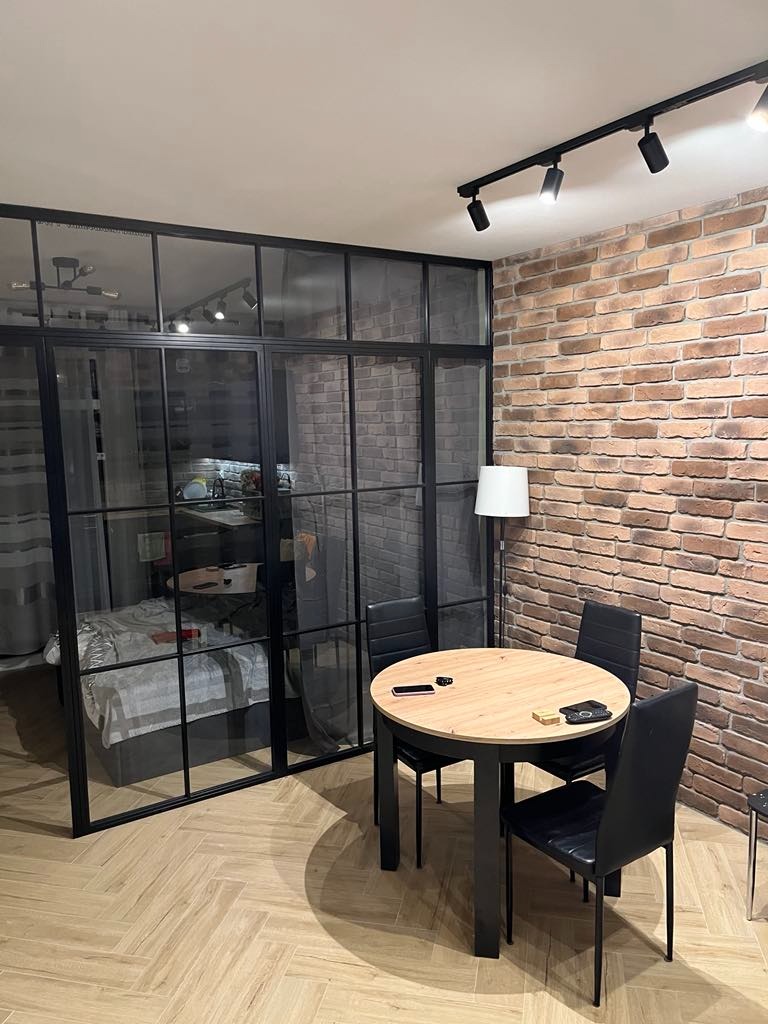Nowoczesne wnętrze z przeszkleniem oddzielającym sypialnię, ceglana ściana i okrągły stół z czarnymi krzesłami na parkiecie ułożonym w jodełkę.