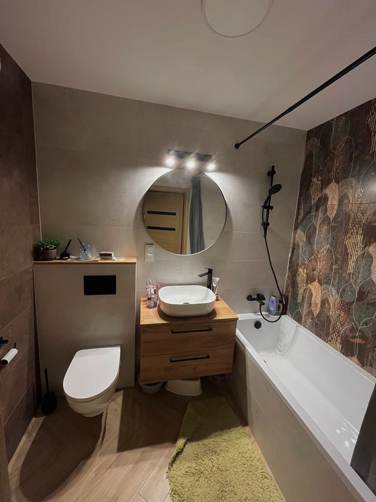 Nowoczesna łazienka z drewnianą szafką pod umywalką, okrągłym lustrem z oświetleniem, białą wanną i toaletą w zabudowie, wykończona płytkami w różnych kolorach i wzorach, z zielonym dywanikiem...