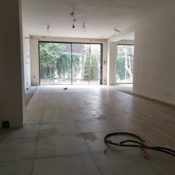 Lutsyk - Wnętrze w trakcie prac wykończeniowych: szare płytki podłogowe, przygotowane ściany do malowania, widoczne przewody elektryczne na podłodze, duże okna z widokiem na zieleń ogrodu, drabina oparta...