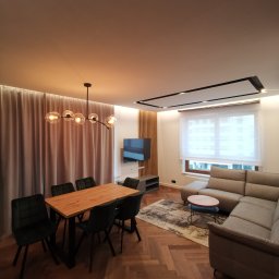 Lutsyk - Wykończone wnętrze salonu z parkietem w jodełkę, szarą sofą narożną, drewnianym stołem z krzesłami i nowoczesną lampą wiszącą z szklanymi kloszami.