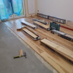 Lutsyk - Układanie jasnych paneli podłogowych, widoczny klej, młotek i paczki paneli marki Barlinek, okno zabezpieczone folią.