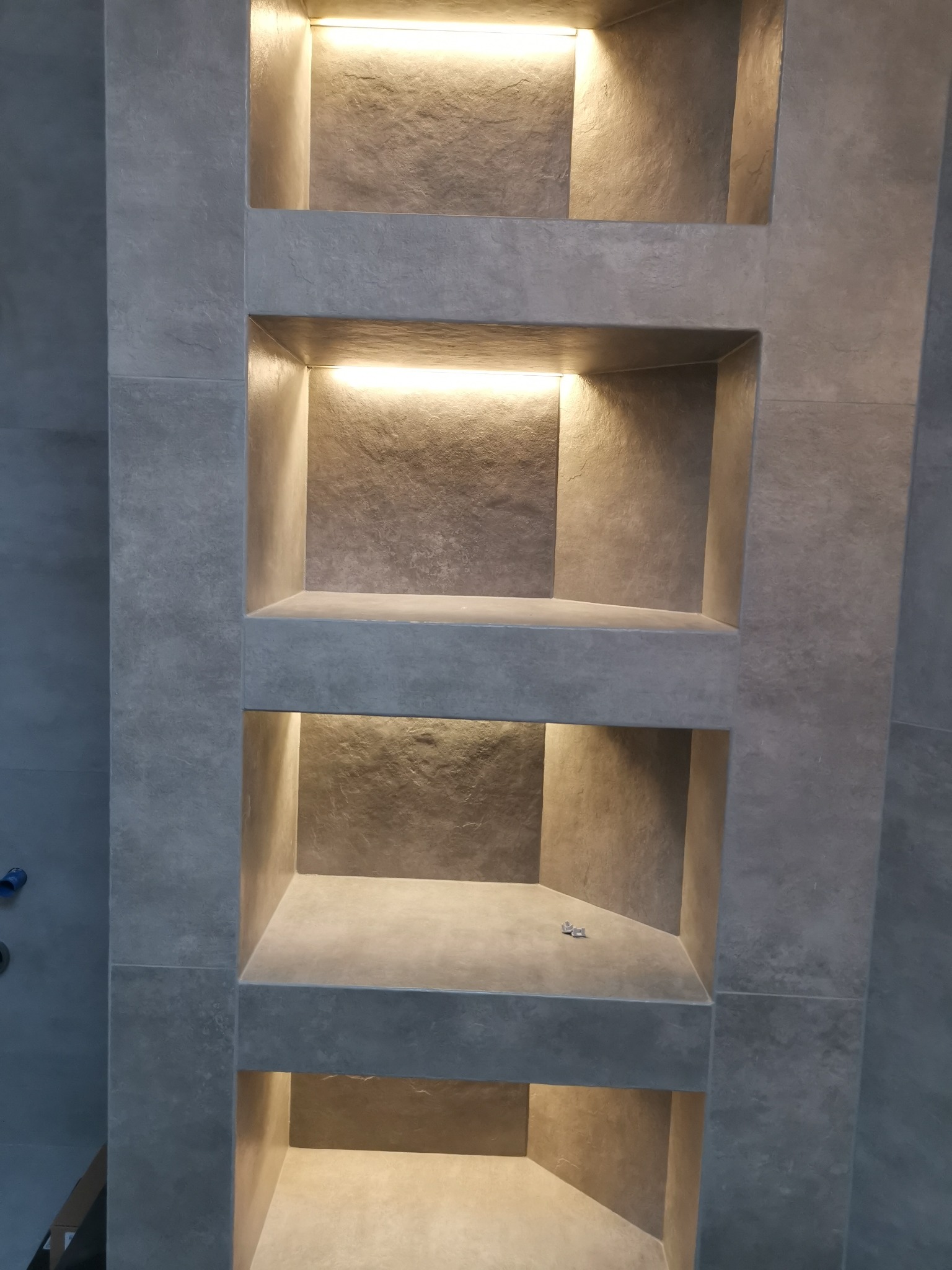 Ściana łazienki wyłożona szarymi płytkami z wnękami oświetlonymi od góry, w jednej z wnęk leżą dwa małe przedmioty.