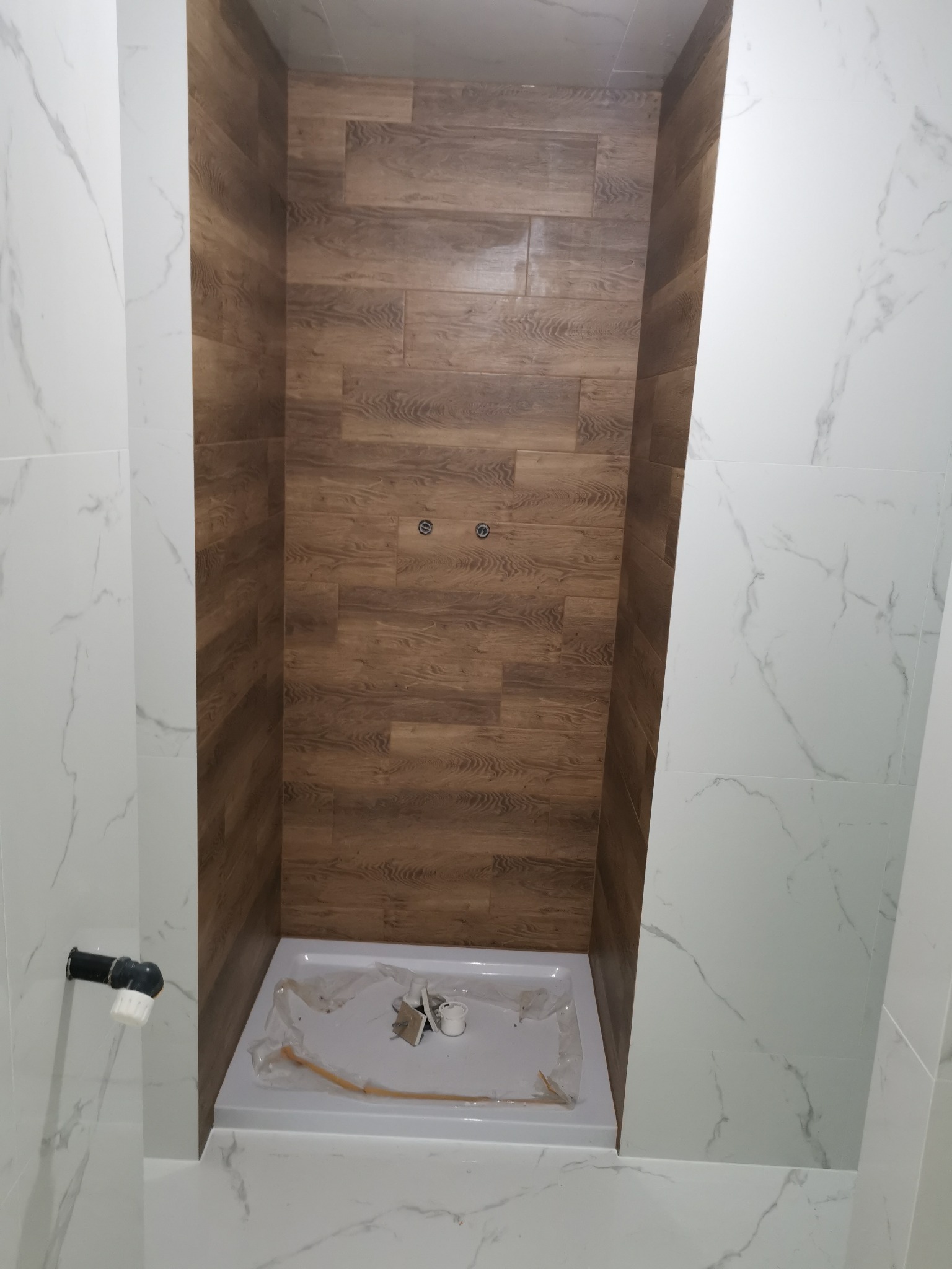 Niedokończona kabina prysznicowa z brodzikiem, wyłożona płytkami imitującymi drewno, otoczona białymi marmurkowymi płytkami ściennymi, widoczne elementy instalacji wodno-kanalizacyjnej.