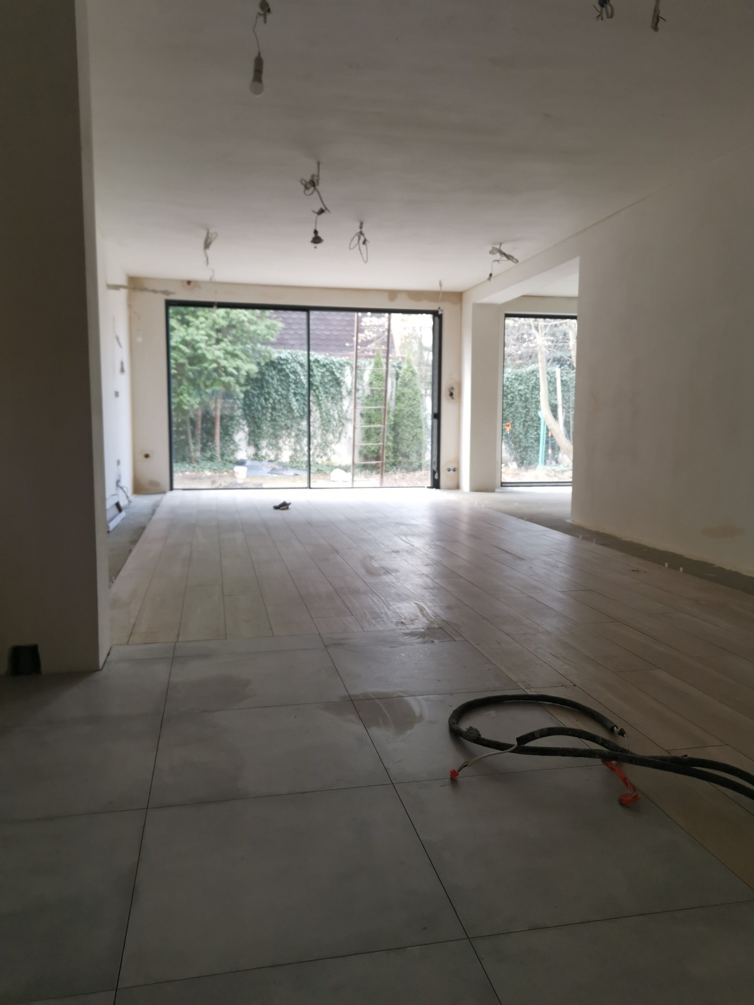 Wnętrze w trakcie prac wykończeniowych: szare płytki podłogowe, przygotowane ściany do malowania, widoczne przewody elektryczne na podłodze, duże okna z widokiem na zieleń ogrodu, drabina oparta...