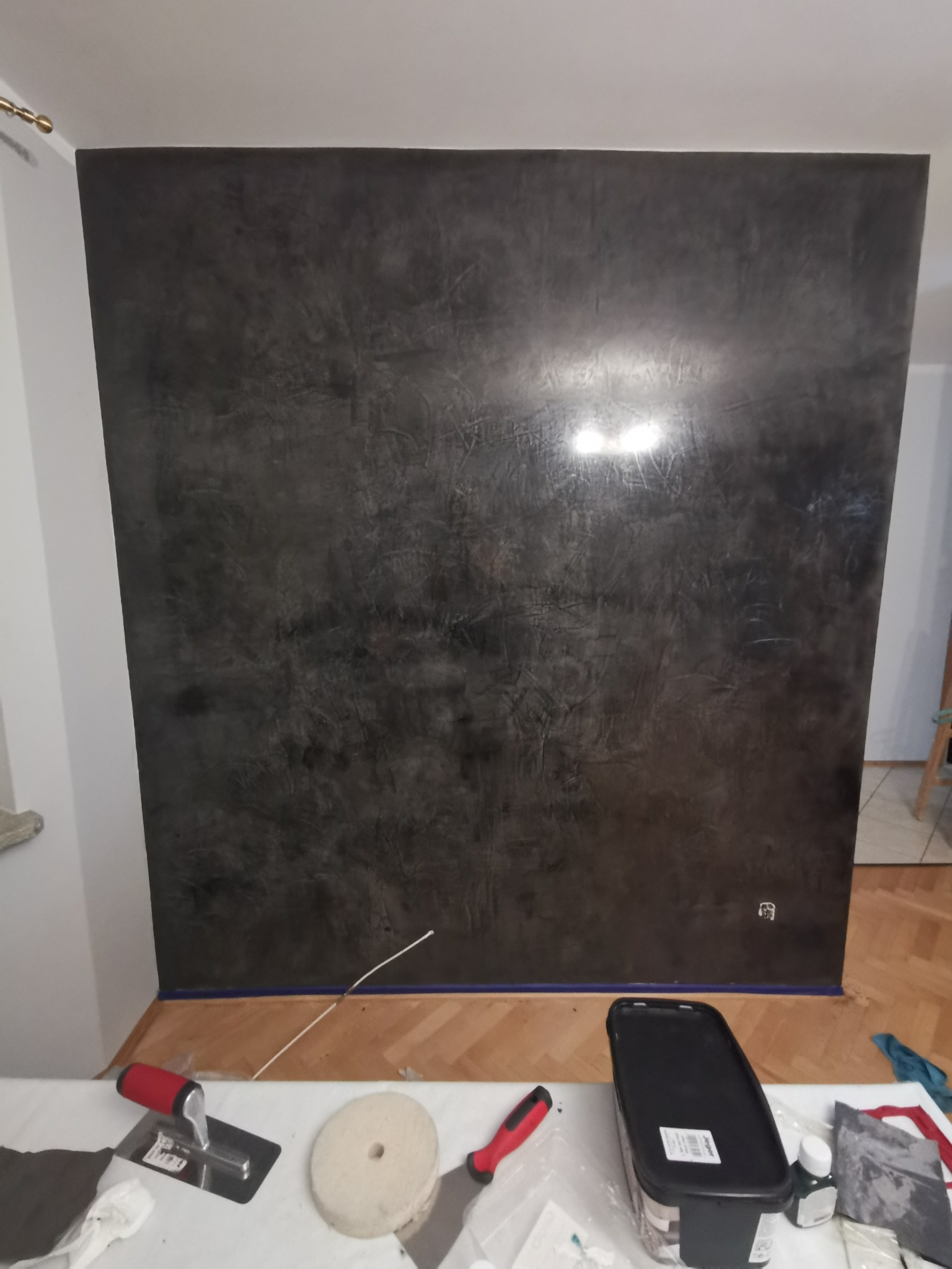 Ściana z efektem dekoracyjnym w odcieniach szarości, zabezpieczona taśmą malarską przy podłodze, z narzędziami i materiałami przygotowanymi do dalszej pracy na blacie.