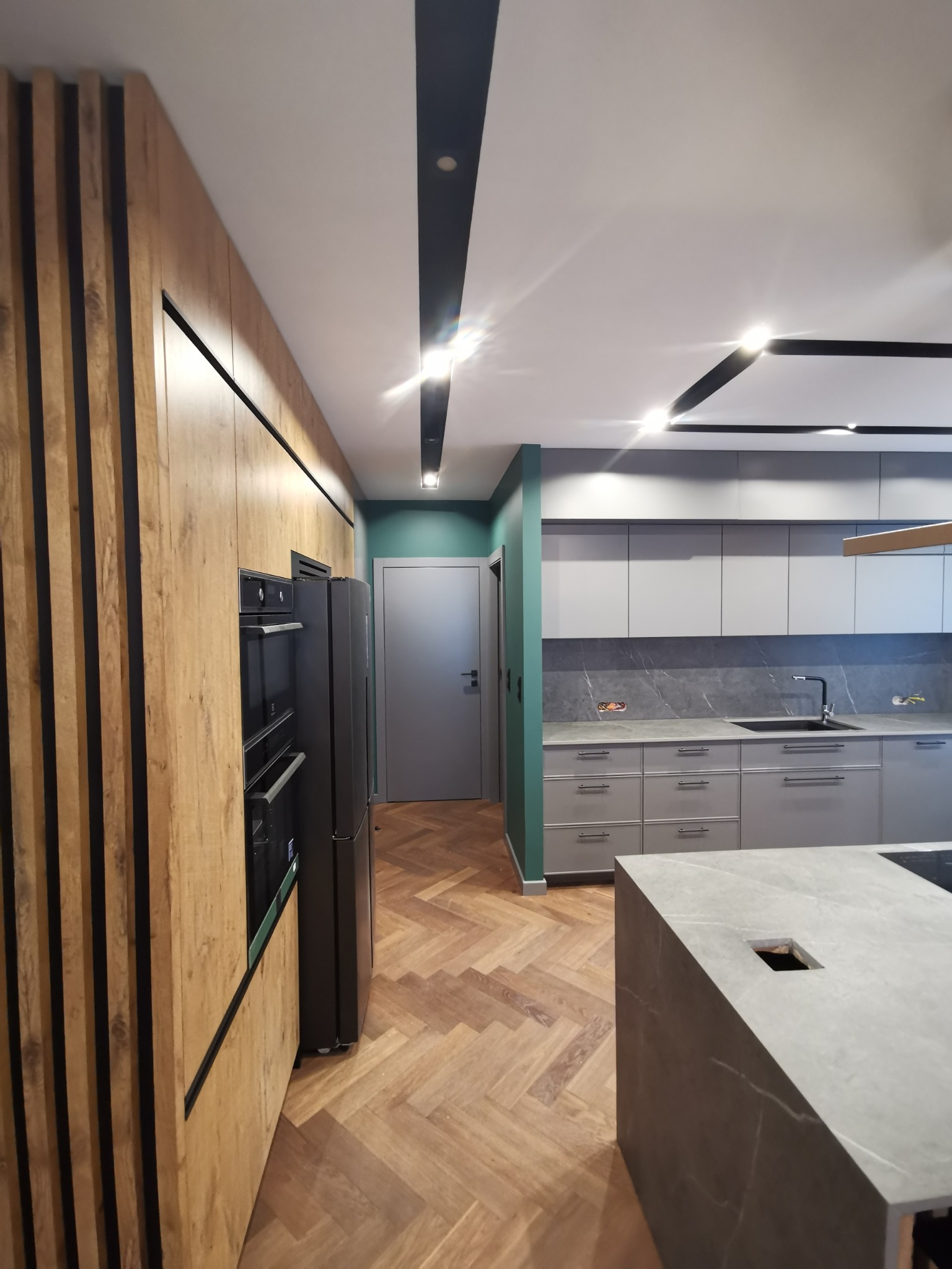 Nowoczesna kuchnia po remoncie: drewniane lamele na zabudowie, szara zabudowa kuchenna z marmurowym backsplash, parkiet w jodełkę, wyspa kuchenna z szarego kamienia.